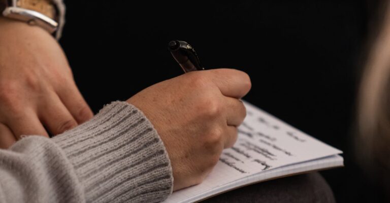 Person in grey sweater taking notes in a notebook with a pen.