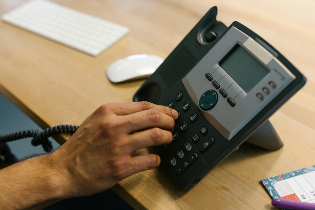 A hand dialing a phone on a wooden desk. Office environment with keyboard and planner.