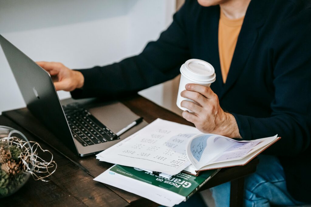 Person working remotely on a laptop with a coffee cup and papers on a wooden table.