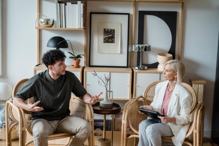 A man and woman in discussion in a modern living room with wooden chairs and artistic decor.