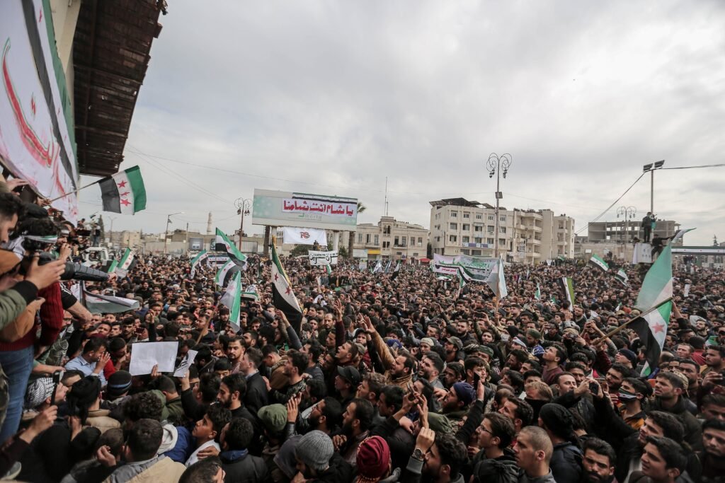 Aerial view of a large political protest in Idlib, Syria, showcasing numerous protesters holding flags and signs.